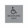 Silvergrå och svart toalettskylt för besökare hwc med piktogram av en rullstol, texten besökare hwc står under symbolen och punktskrift längst ned