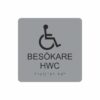 Silvergrå och svart toalettskylt för besökare hwc med piktogram av en rullstol, texten besökare hwc står under symbolen och punktskrift längst ned